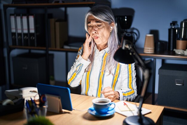 Mulher de meia idade com cabelos grisalhos trabalhando no escritório à noite tocando a boca com a mão com expressão dolorosa por causa de dor de dente ou doença dentária no dentista de dentes