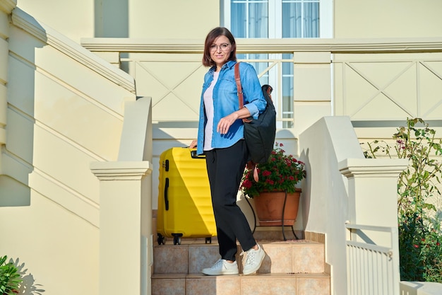 Mulher de meia idade com mochila com mala nos degraus da varanda da casa