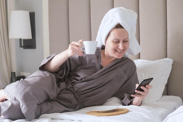 Mulher de meia idade com toalha na cabeça está deitada na cama sorrindo e olhando para a tela do smartphone