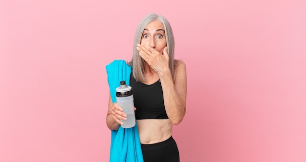 Mulher de meia-idade de cabelo branco cobrindo a boca com as mãos com uma toalha chocada e uma garrafa de água. conceito de fitness