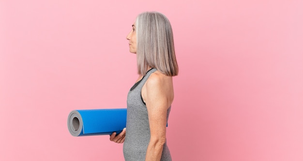 Mulher de meia-idade de cabelo branco em vista de perfil, pensando, imaginando ou sonhando acordada e segurando um tapete de ioga. conceito de fitness