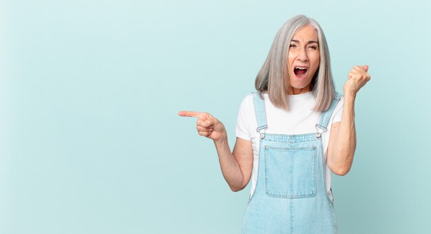 Mulher de meia-idade de cabelo branco gritando agressivamente com uma expressão de raiva e apontando para o lado