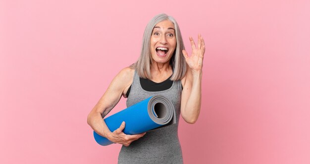 Mulher de meia-idade de cabelo branco gritando com as mãos no ar e segurando um tapete de ioga. conceito de fitness