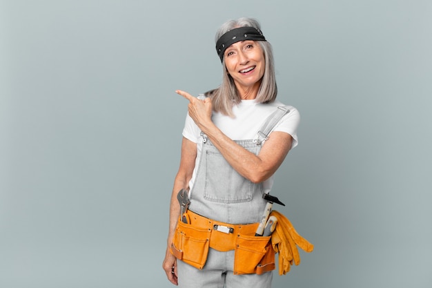 Mulher de meia-idade de cabelo branco parecendo animada e surpresa, apontando para o lado e usando roupas de trabalho e ferramentas. conceito de limpeza