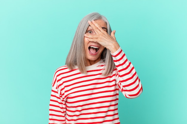 Mulher de meia-idade de cabelo branco parecendo chocada, assustada ou apavorada, cobrindo o rosto com a mão