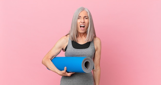 Mulher de meia-idade de cabelo branco parecendo muito chocada ou surpresa e segurando um tapete de ioga. conceito de fitness