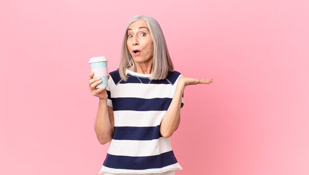 Mulher de meia-idade de cabelo branco parecendo surpresa e chocada, com o queixo caído segurando um objeto e segurando uma embalagem de café para viagem