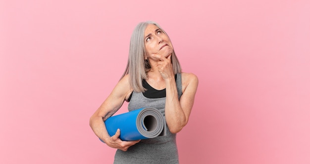 Mulher de meia-idade de cabelo branco pensando, sentindo-se duvidosa e confusa e segurando um tapete de ioga. conceito de fitness