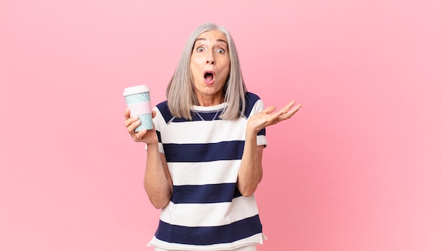 Mulher de meia-idade, de cabelo branco, sentindo-se extremamente chocada e surpresa, segurando um recipiente para levar café