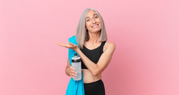 Mulher de meia-idade de cabelo branco sorrindo alegremente, sentindo-se feliz e mostrando um conceito com uma toalha e uma garrafa de água. conceito de fitness