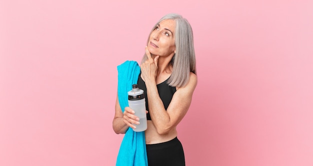 Mulher de meia-idade de cabelo branco sorrindo com uma expressão feliz e confiante com a mão no queixo com uma toalha e uma garrafa de água. conceito de fitness