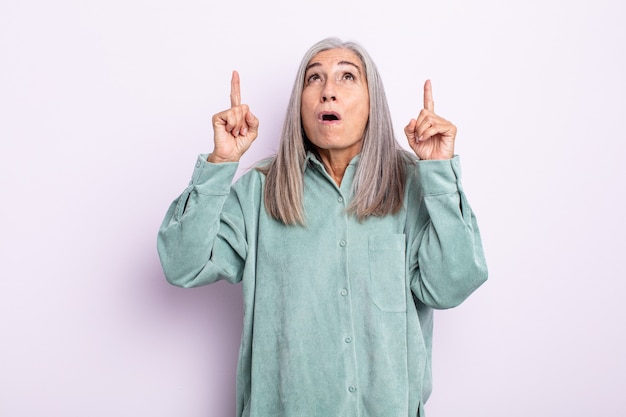 Mulher de meia-idade de cabelo grisalho parecendo chocada, surpresa e boquiaberta, apontando para cima com as duas mãos para copiar o espaço