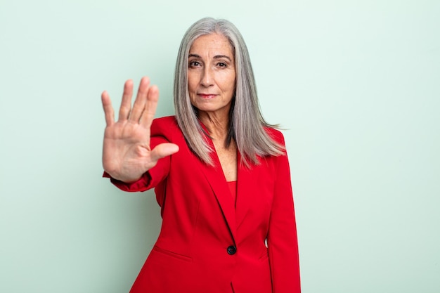 Mulher de meia-idade de cabelos grisalhos olhando sério, mostrando a palma da mão aberta, fazendo gesto de parada. conceito de mulher de negócios sênior