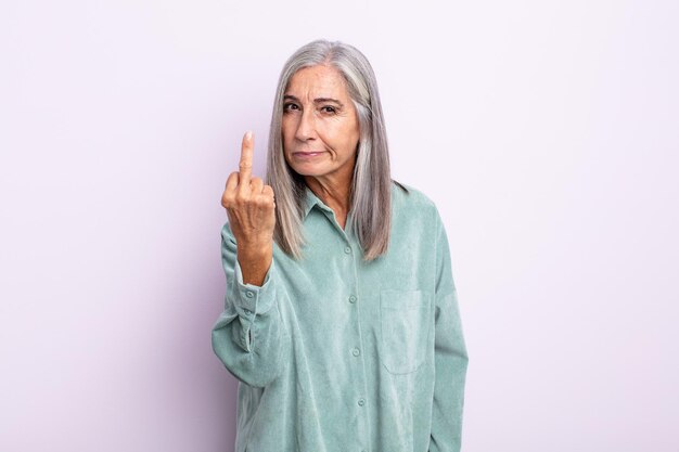 Mulher de meia-idade de cabelos grisalhos se sentindo zangada, irritada, rebelde e agressiva, sacudindo o dedo do meio, lutando contra