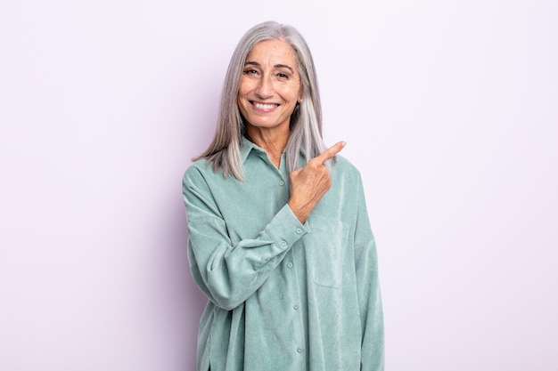 Mulher de meia-idade de cabelos grisalhos sorrindo alegremente, sentindo-se feliz e apontando para o lado e para cima, mostrando o objeto no espaço da cópia