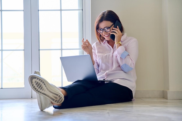 Mulher de meia idade de negócios sentada no chão com um laptop falando em um telefone celular