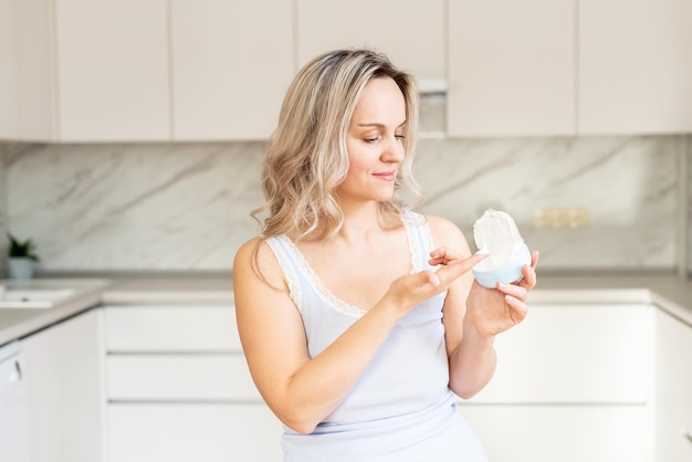 Mulher de meia idade de pijama na cozinha com creme hidratante Cuidados faciais Cosmetologia da pele Rotina matinal Spa em casa e procedimentos cosméticos de bem-estar tratamento da pele do rosto