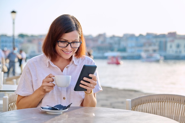 Mulher de meia idade descansando no café ao ar livre com uma xícara de café usando um smartphone