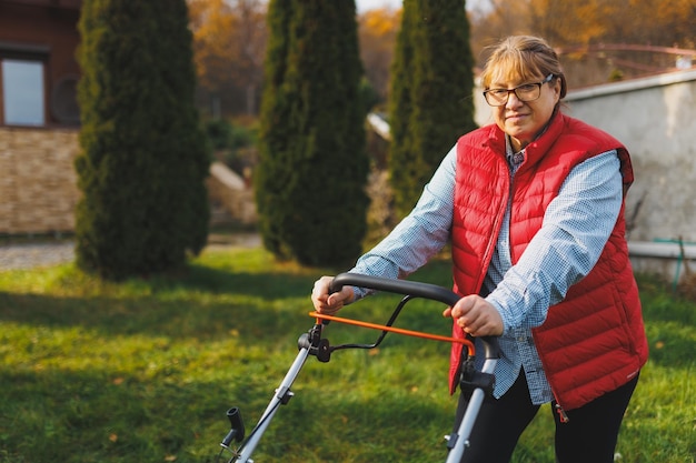 Mulher de meia idade em colete vermelho usando cortador de grama no quintal olhando para a câmera Jardineira feminina trabalhando no verão ou outono cortando grama no quintal Conceito de natureza de trabalho de jardinagem