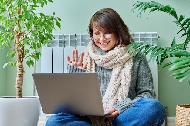 Mulher de meia-idade em roupas quentes perto do radiador de aquecimento usando laptop para videochamada