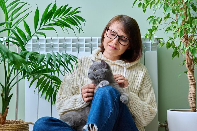 Foto mulher de meia-idade em suéter de lã de tricô com gato sentado perto do radiador de aquecimento animais de estimação sentados nos braços da mulher proprietária e gato aquecendo juntos temporada inverno outono aquecimento estilo de vida de lazer economia de energia