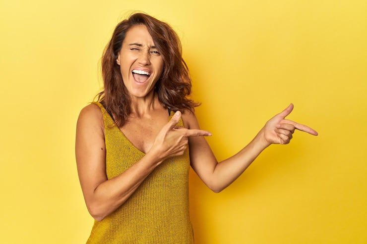 Foto mulher de meia idade em um pano de fundo amarelo apontando com os dedos indicadores para um espaço de cópia