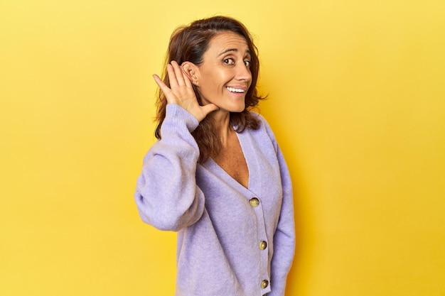 Mulher de meia idade em um pano de fundo amarelo tentando ouvir uma fofoca
