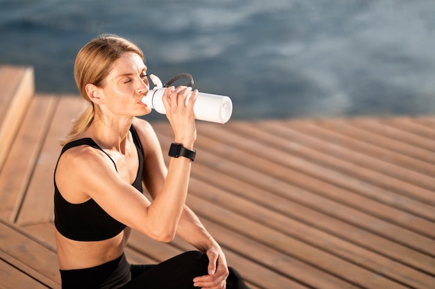 Mulher de meia-idade esportiva cansada bebendo água da garrafa após o treino ao ar livre