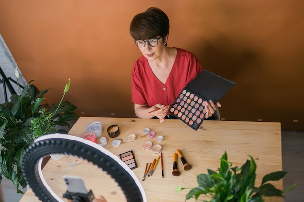 Foto mulher de meia idade falando sobre cosméticos com sombras de maquiagem e paleta de blush e pincéis enquanto grava seu vídeo mulher madura fazendo vídeo para seu blog sobre cosméticos