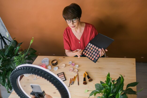 Mulher de meia idade falando sobre cosméticos com sombras de maquiagem e paleta de blush e pincéis