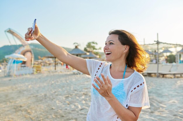 Mulher de meia idade feliz falando no smartphone usando videochamada na praia