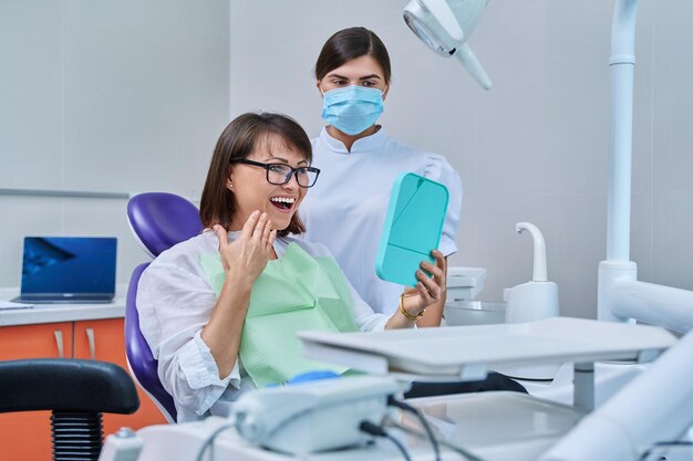 Mulher de meia-idade feliz junto com o paciente do dentista sentado na cadeira dentária olhando para o espelho Tratamento de próteses Implantação de dentes dentários conceito de cuidados de beleza de saúde