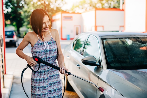 Mulher de meia-idade feliz lavando carro na estação de lavagem de carros usando máquina de água de alta pressão.