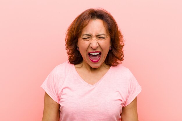 Mulher de meia idade gritando agressivamente, parecendo muito irritada, frustrada, indignada ou irritada, gritando não contra a parede rosa