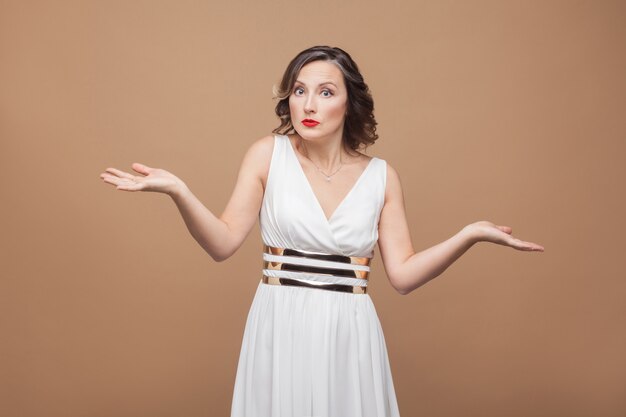 Mulher de meia-idade intrigada diz que não sei. Mulher de expressão emocional em vestido branco, maquiagem, lábios vermelhos e penteado encaracolado escuro. Foto de estúdio, interna, isolada em um fundo bege ou marrom claro