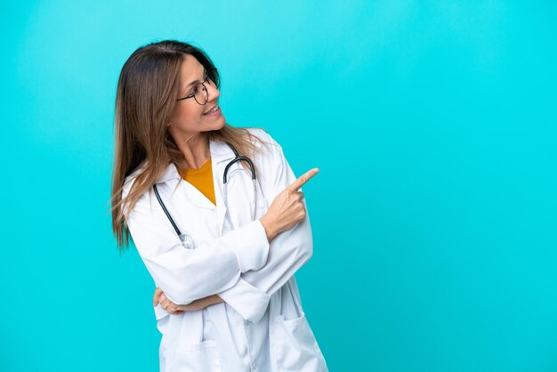 Mulher de meia idade isolada em fundo azul, vestindo um vestido de médico e apontando o lado