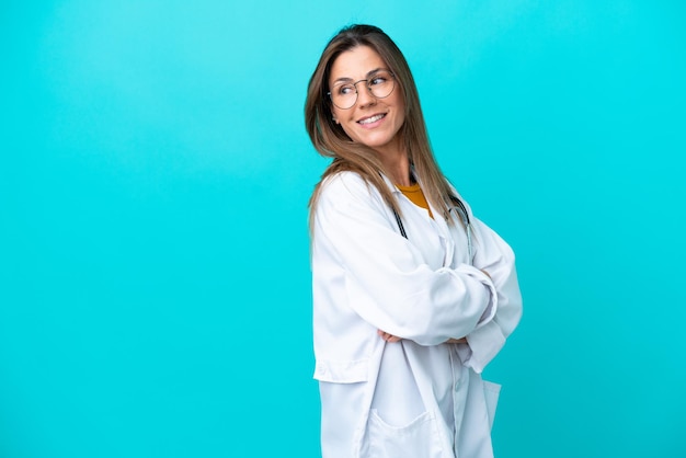 Mulher de meia idade isolada em fundo azul, vestindo um vestido de médico e com os braços cruzados