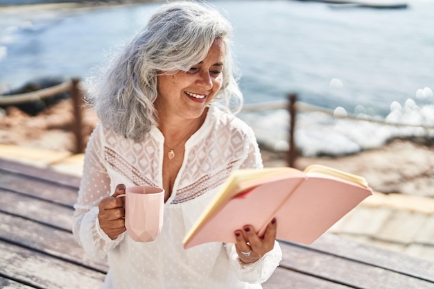 Mulher de meia-idade lendo livro e tomando café sentado no banco à beira-mar