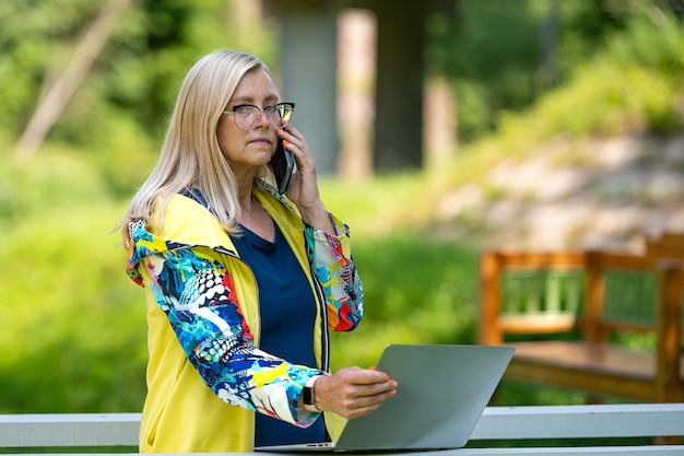 Mulher de meia idade no jardim trabalhando em casa usando laptop e falando ao telefone