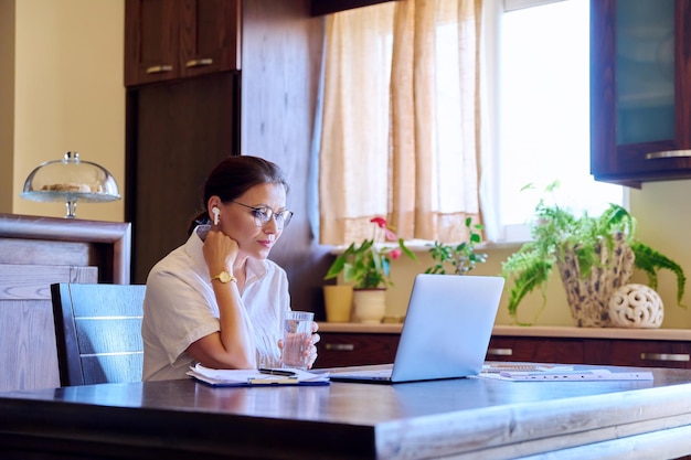 Mulher de meia idade no local de trabalho doméstico em fones de ouvido trabalhando usando laptop
