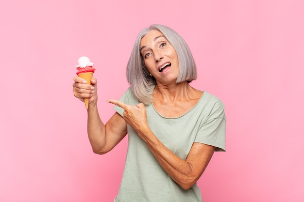 Mulher de meia-idade parecendo animada e surpresa apontando para o lado e para cima para copiar o espaço tomando um sorvete