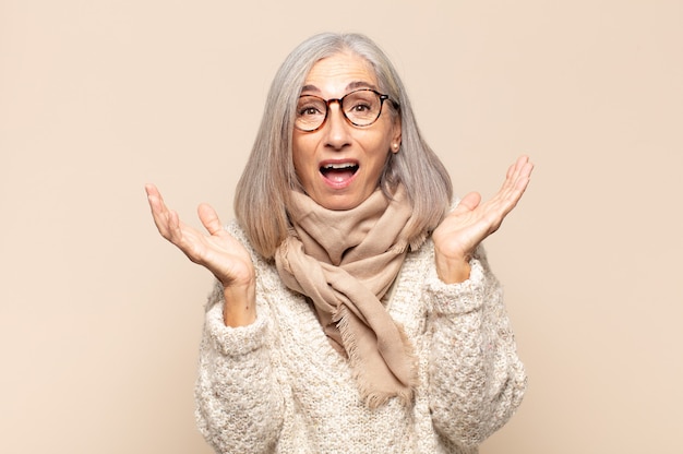 Mulher de meia-idade parecendo feliz e animada, chocada com uma surpresa inesperada com as duas mãos abertas ao lado do rosto