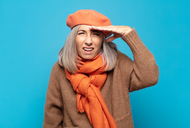 Mulher de meia-idade parecendo perplexa e atônita, com a mão na testa olhando para longe, observando ou procurando