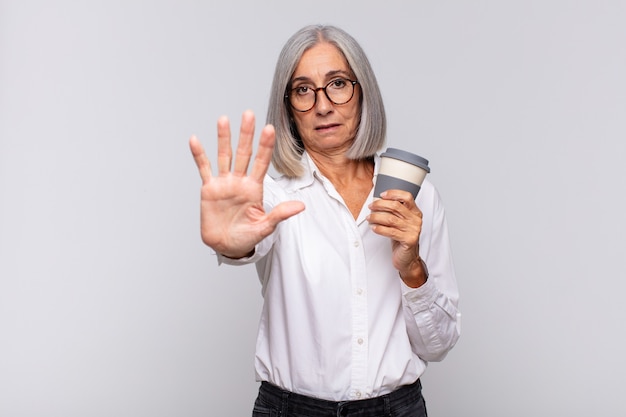 Mulher de meia-idade parecendo séria, severa, descontente e irritada mostrando a palma da mão aberta fazendo gesto de parada conceito de café