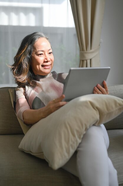 Mulher de meia idade positiva navegando na Internet sem fio no tablet relaxa no sofá em casa Conceito de tecnologia idosa