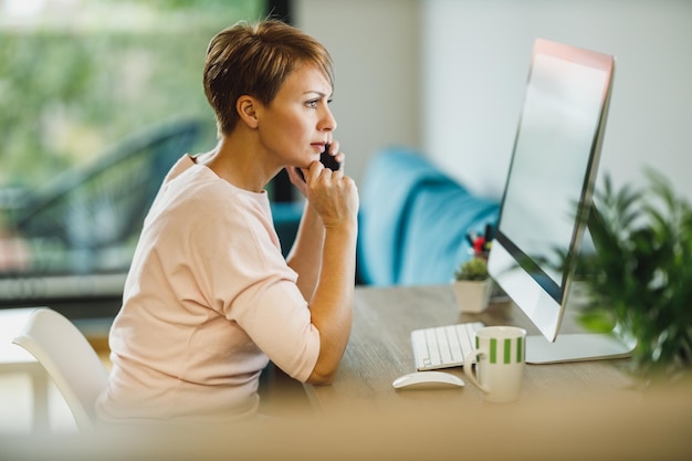 Mulher de meia idade preocupada falando em um smartphone enquanto trabalhava no computador em casa.