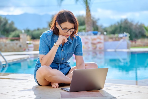 Mulher de meia idade relaxando perto da piscina usando um laptop