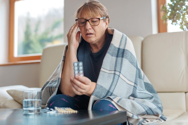 Mulher de meia idade segura uma bolha de comprimidos e lê instruções médicas sentada no sofá
