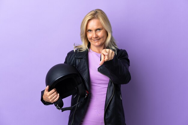 Mulher de meia-idade segurando um capacete de motociclista isolado em roxo surpresa e apontando para a frente