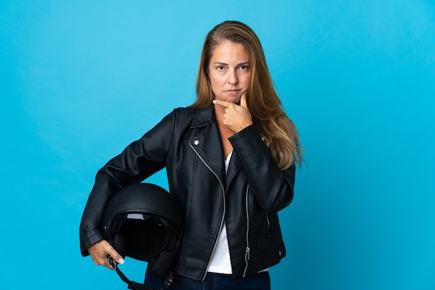 Mulher de meia-idade segurando um capacete de motociclista isolado na parede azul pensando